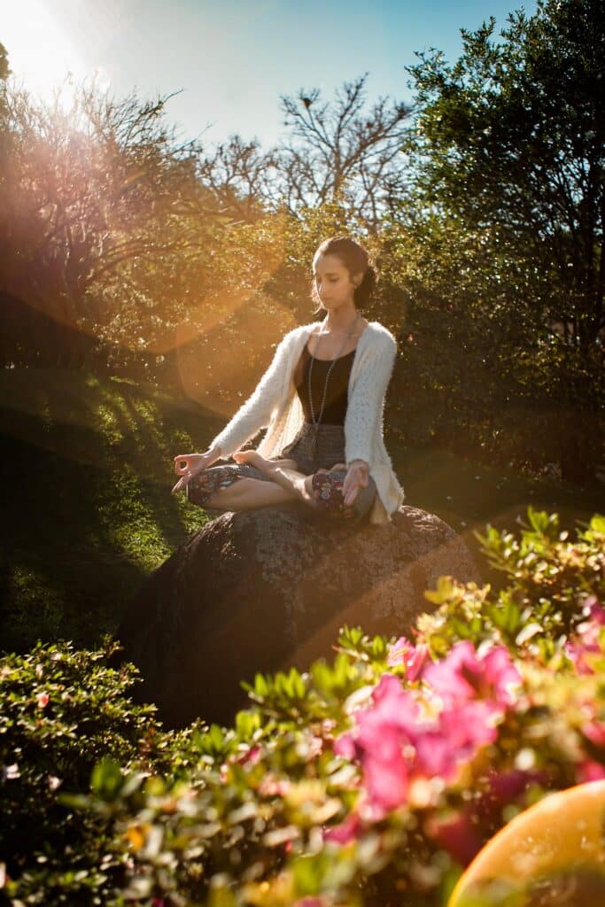 woman meditating in sunlight on a rock in a garden, surrounded by pink flowers and green bushes, with a calm expression and eyes closed