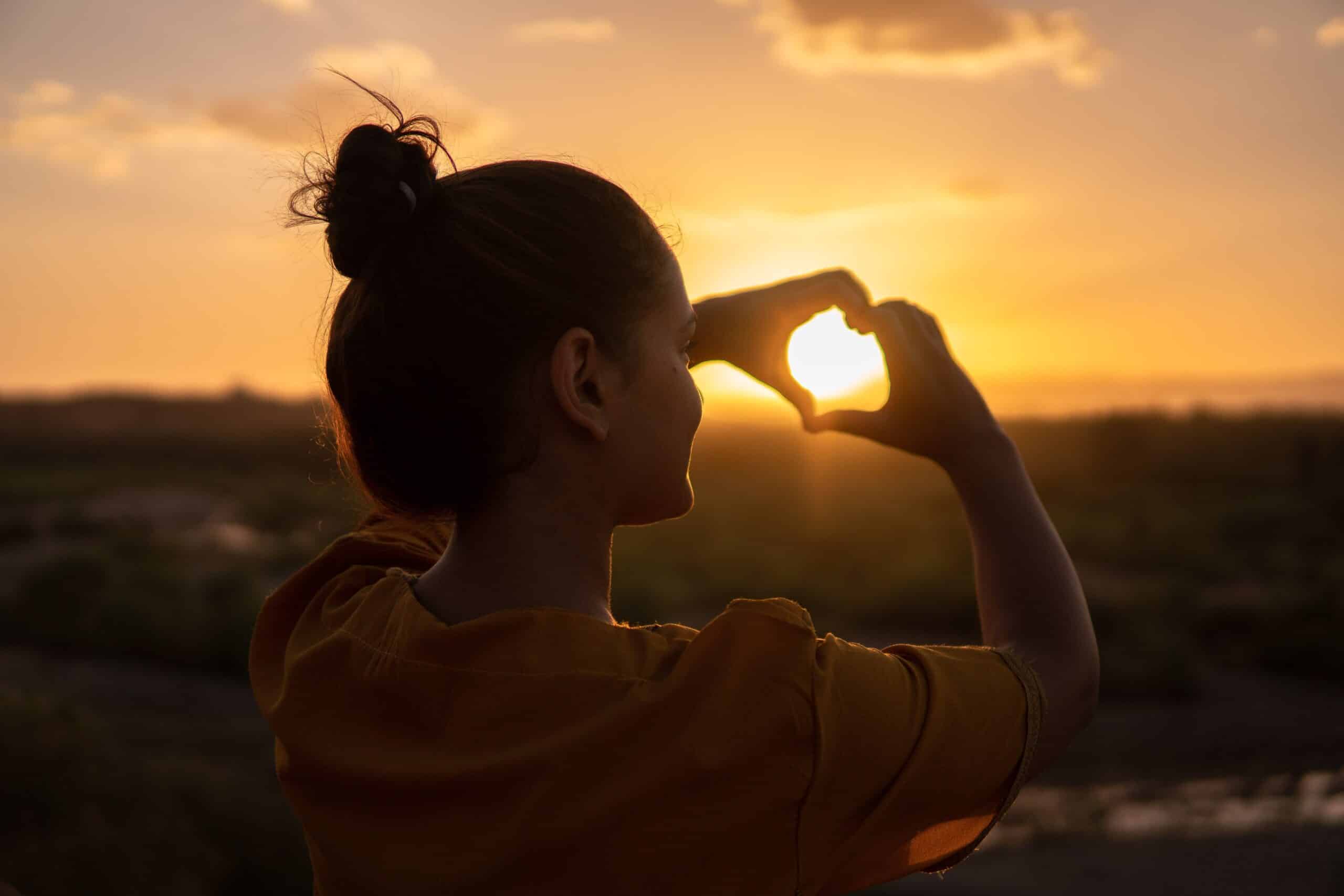 Silhouette of a woman creating a heart shape with her hands around the setting sun, against a warm sunset sky