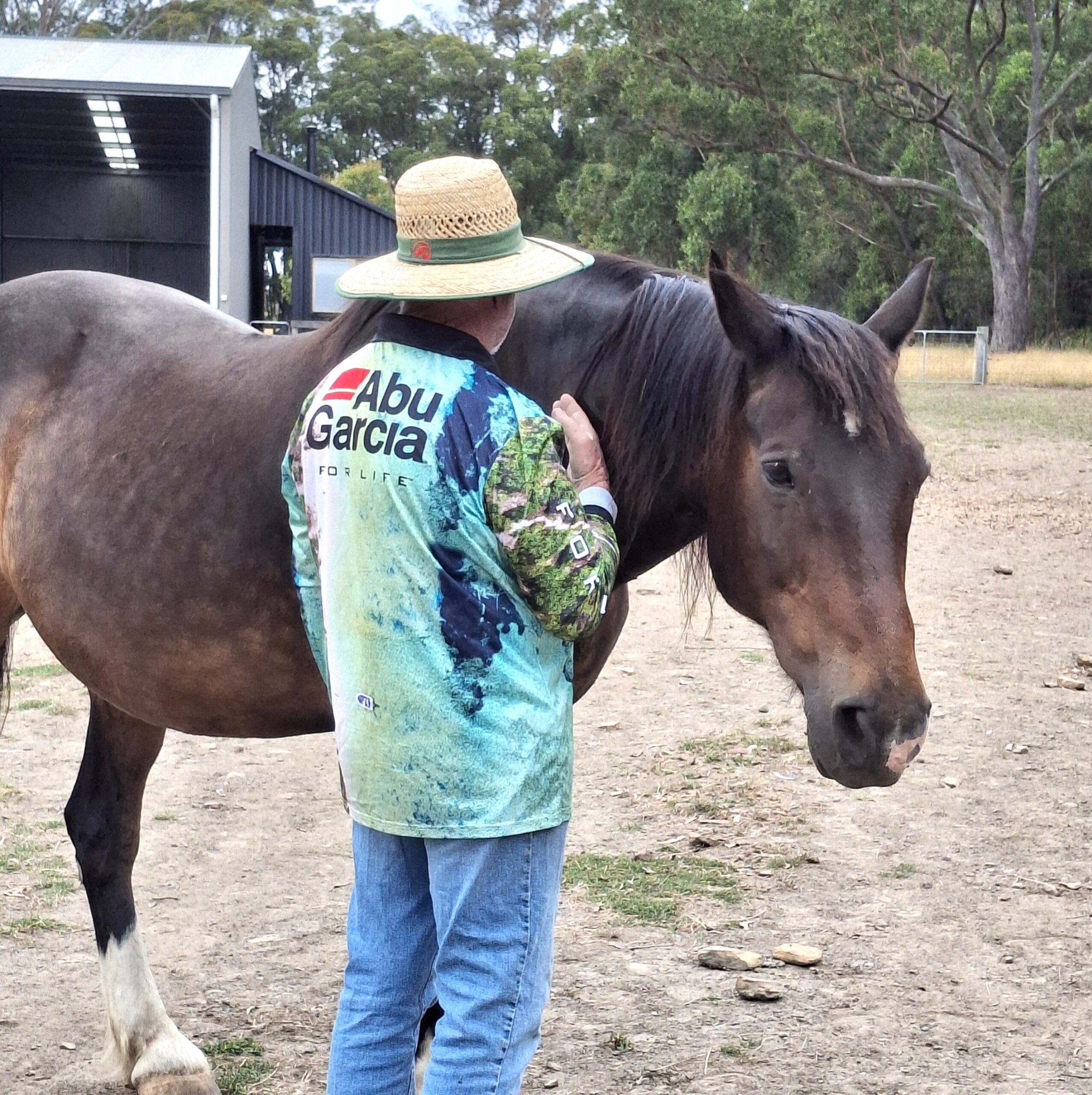 A person wearing a wide-brimmed hat and an Abu Garcia shirt stands beside a brown horse, gently touching its neck in an outdoor rural area with trees and a shed in the background.