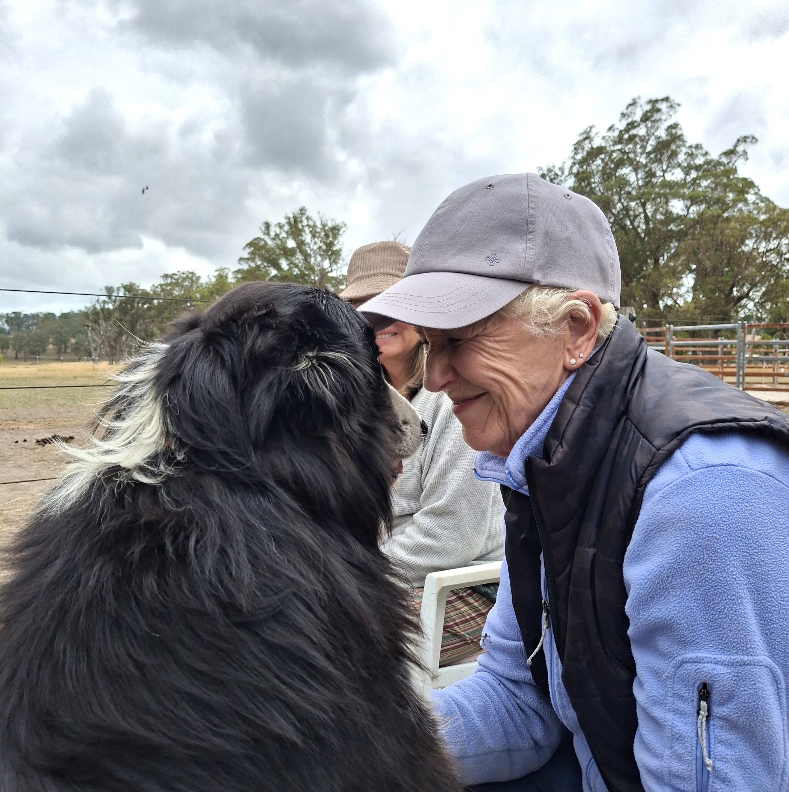 An elderly woman in a gray cap and blue jacket smiles warmly at a black and white dog, sitting close together outdoors. Another person sits in the background, with trees and cloudy skies behind them.