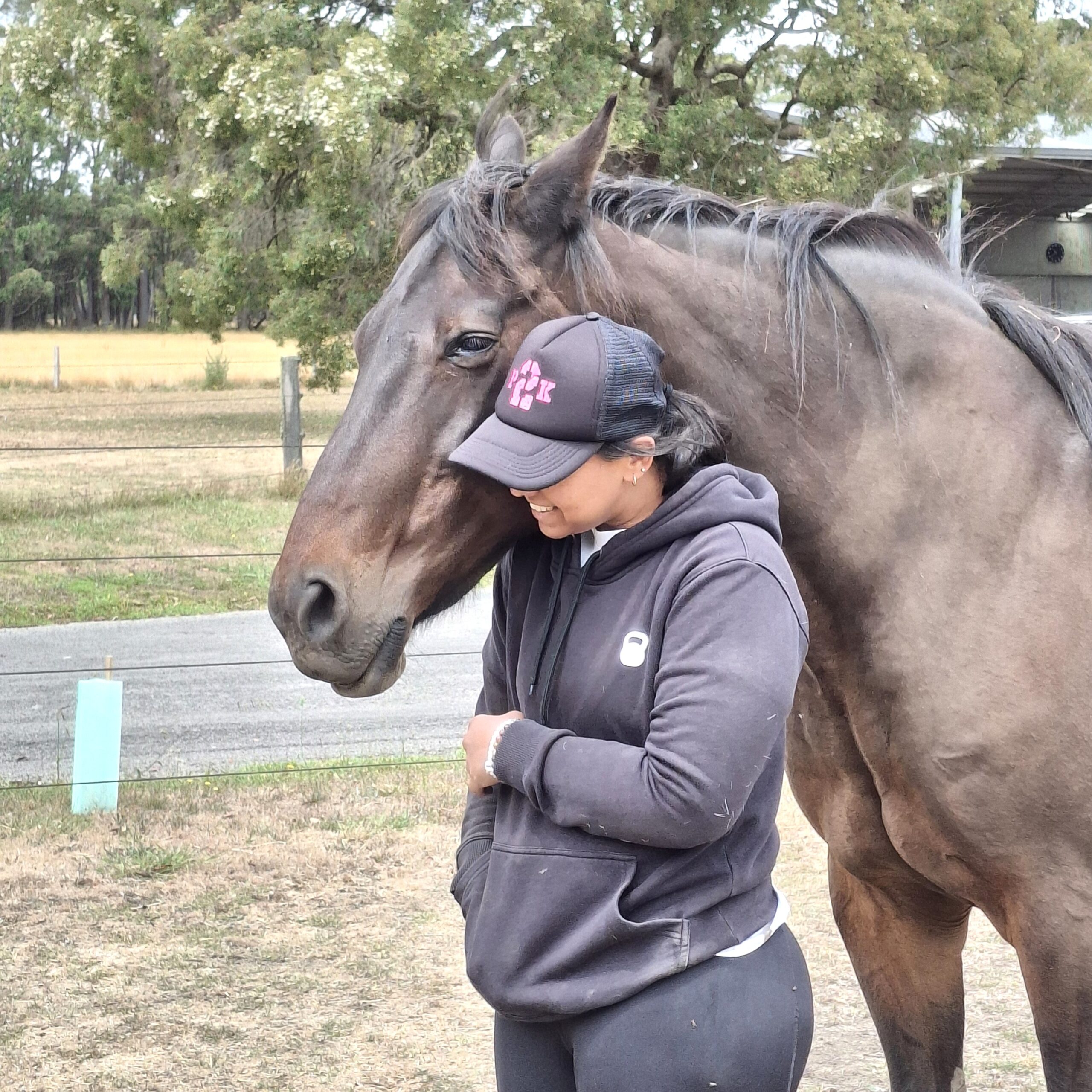 A person in a black hoodie and cap stands beside a brown horse, smiling as the horse gently rests its head on their shoulder in an outdoor rural setting with trees and grass.