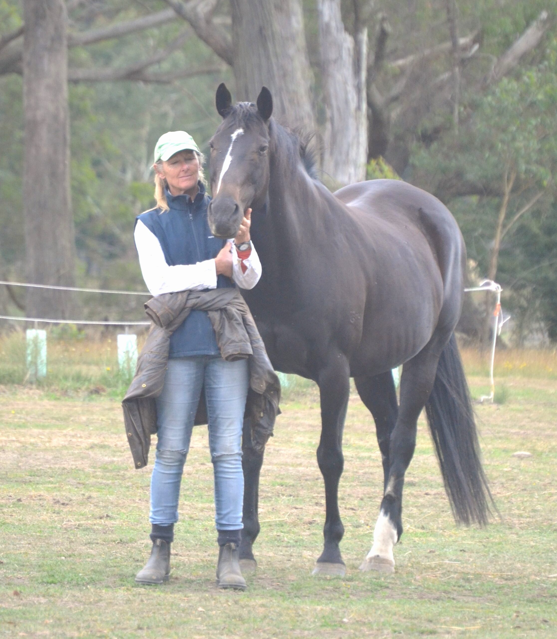 soo murray 1 A woman wearing a green cap, blue vest, and jeans stands beside a dark brown horse with a white stripe on its face. They are outdoors on grass, with trees and fencing in the background.