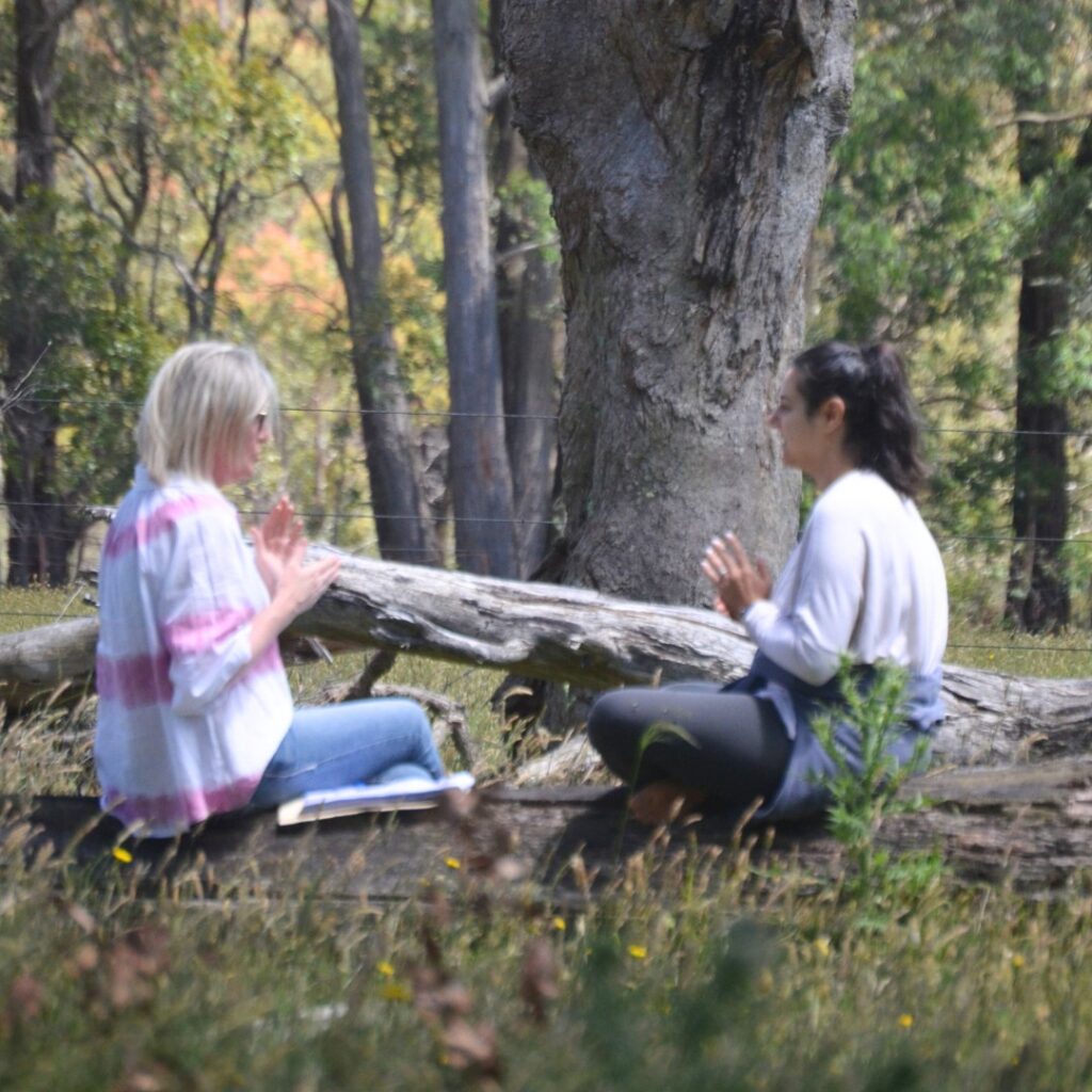 Two women sit facing each other outdoors on a fallen log in a wooded area, appearing to engage in a mindful or meditative activity with hands raised. Trees and greenery surround them.